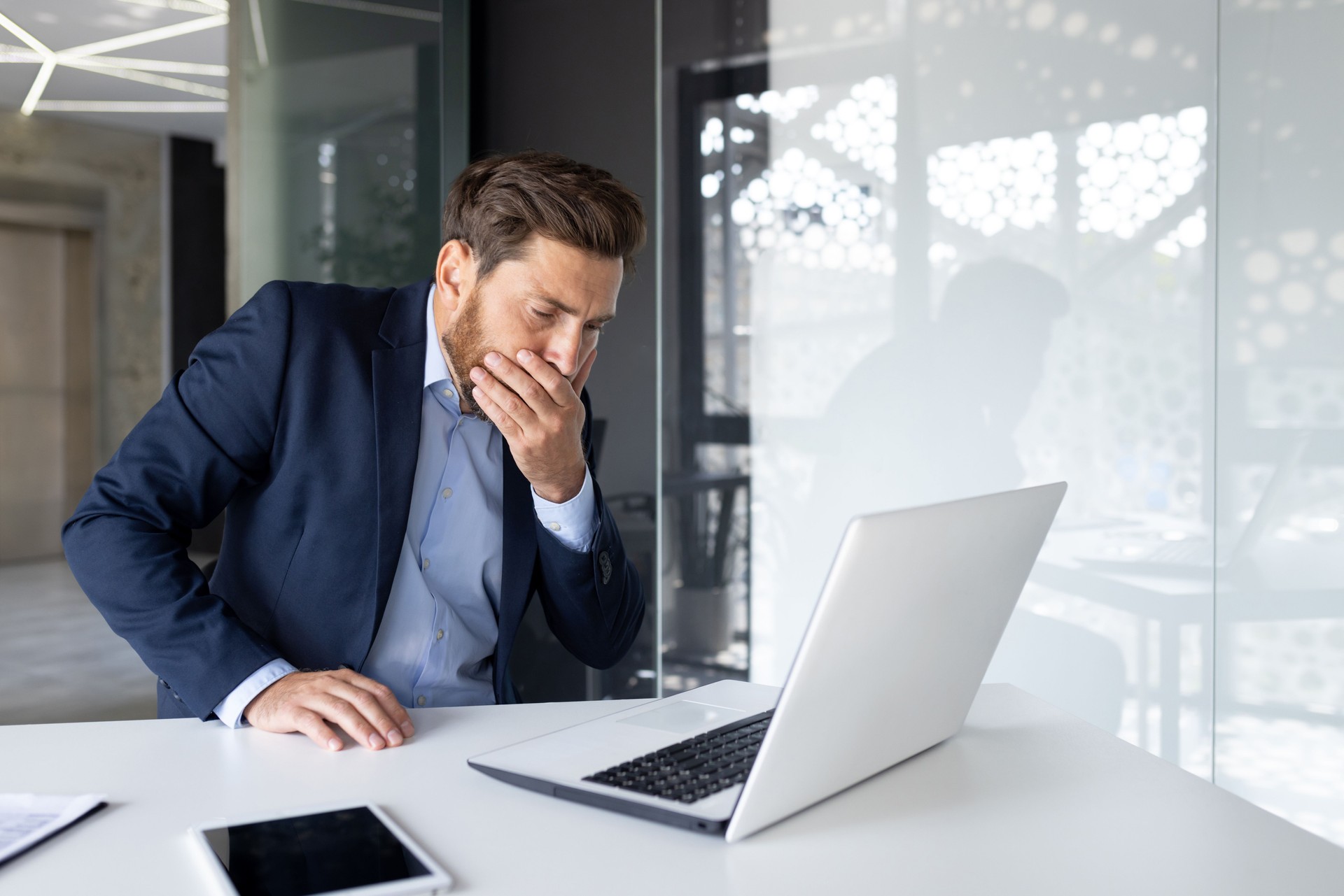 A young man businessman sits in the office at the work table and covers his mouth from nausea, he does not feel well